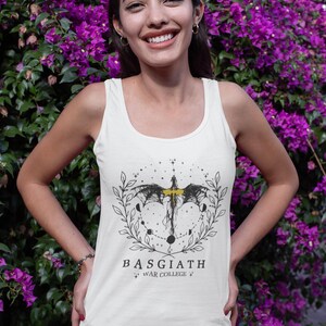 May include: Cream tank top with a black and yellow graphic design. The design features a bat-like creature, moons, and a laurel wreath, with the text "BASGIATH WAR COLLEGE". The tank top is set against a backdrop of purple flowers.