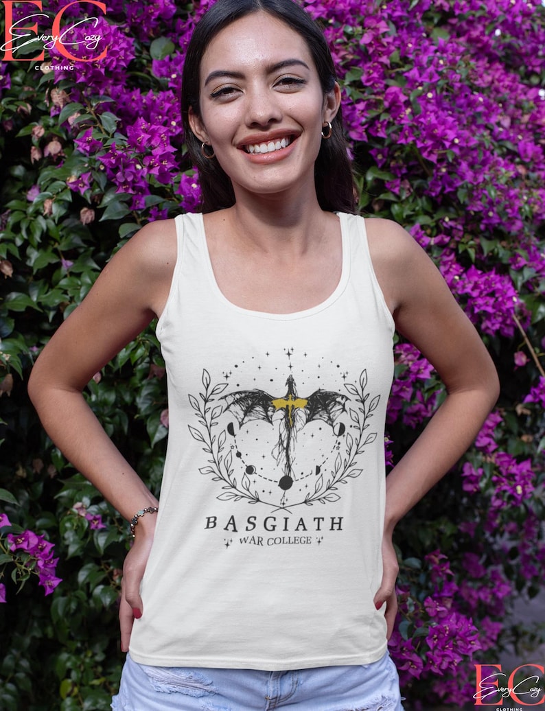 May include: Cream tank top with a black and yellow graphic design. The design features a bat-like creature, moons, and a laurel wreath, with the text "BASGIATH WAR COLLEGE". The tank top is set against a backdrop of purple flowers.