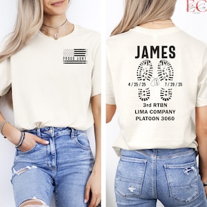 May include: Cream-colored t-shirt with the text "PROUD AUNT" and a black and white American flag graphic on the front. The back of the shirt features the name "JAMES", boot prints, and military unit information.