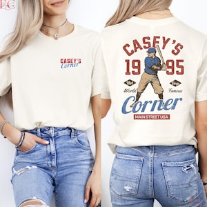May include: Cream-colored t-shirt with "Casey's Corner" printed on the front and back. The back features a baseball player graphic with the text "Casey's 1995 Corner, Main Street USA". The shirt has short sleeves and a crew neck.