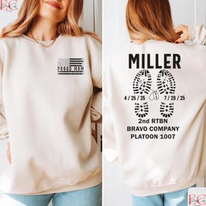 May include: Cream-colored sweatshirt with a black graphic of an American flag and the words "PROUD MOM." The back features the name "MILLER" above two boot prints, dates, and military unit information. A comfortable, casual top.