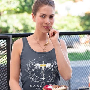 May include: A dark grey vest top with a white graphic design featuring a winged creature, moons, and the text "BASGIATH + WAR CO". The top is worn by a person. A bowl of porridge with berries is on the table.