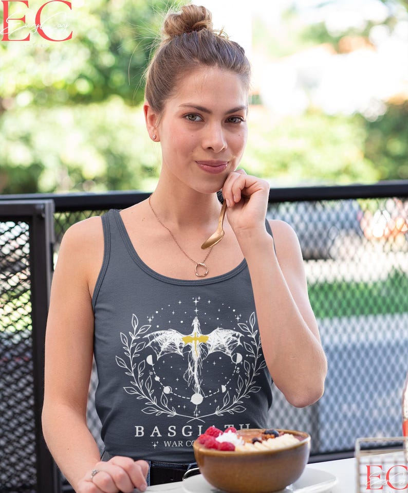 May include: A dark grey vest top with a white graphic design featuring a winged creature, moons, and the text "BASGIATH + WAR CO". The top is worn by a person. A bowl of porridge with berries is on the table.