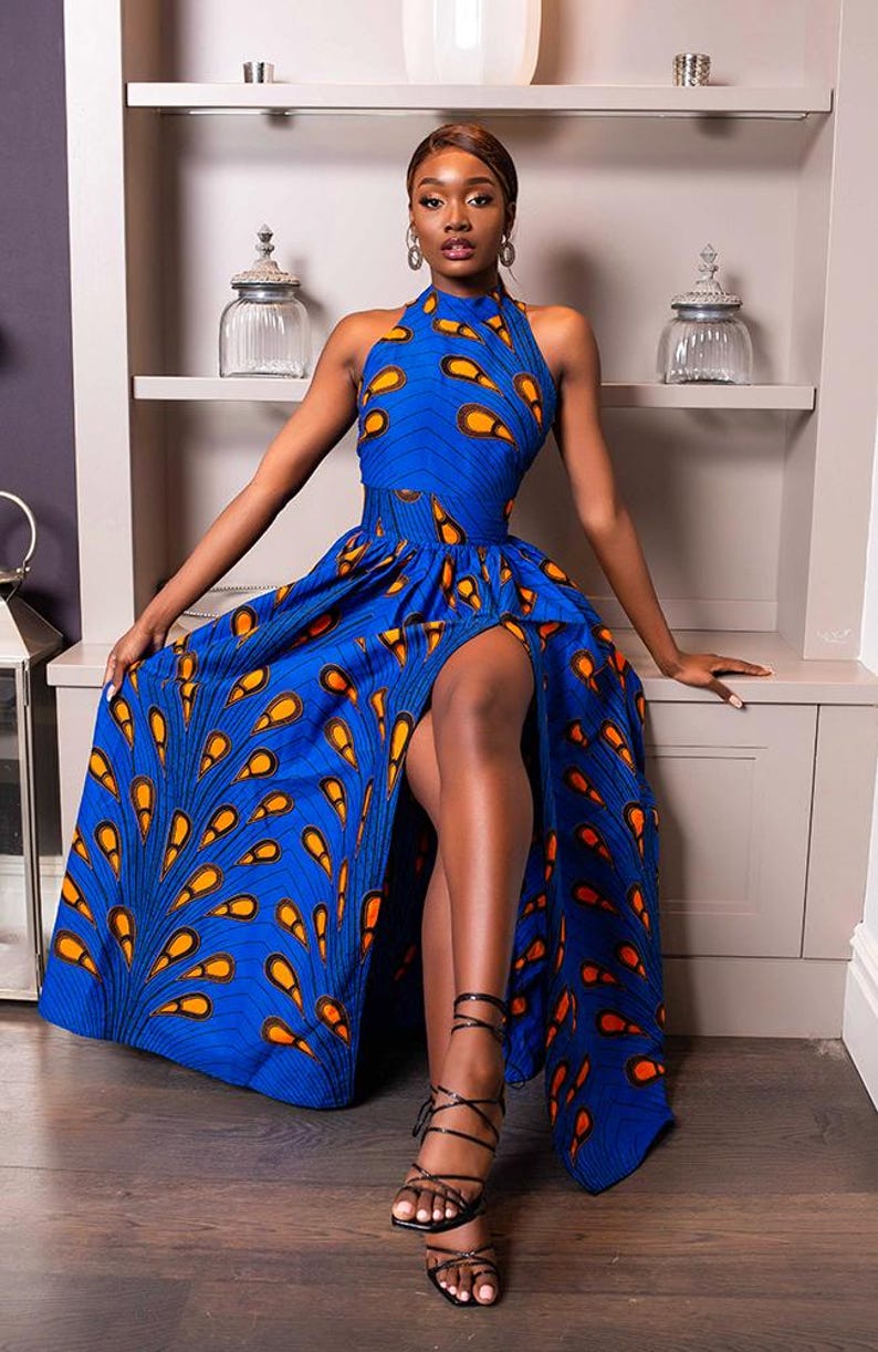 May include: A floor-length sleeveless dress in a vibrant blue fabric featuring a repeating orange and black teardrop pattern. The garment has a high halter neckline, a fitted waist, and a dramatic front thigh-high slit in the full skirt.