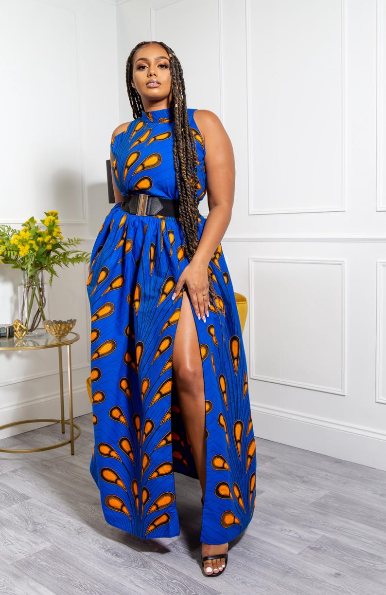 May include: A sleeveless maxi dress in a vibrant blue fabric featuring a repeating orange and black teardrop pattern. The dress has a high neckline, a fitted waist accented by a wide black belt with a gold buckle, and a high front leg slit.