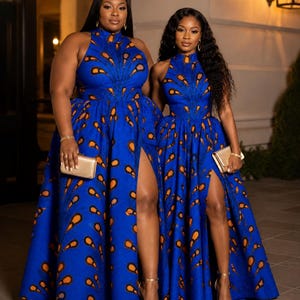 May include: Two floor-length blue dresses featuring a high neckline and a repeating orange and black teardrop pattern. Each dress has a fitted bodice, a full skirt with a high side slit, and is paired with a small gold clutch and gold sandals.