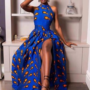 May include: A floor-length sleeveless dress in a vibrant blue fabric featuring a repeating orange and black teardrop pattern. The garment has a high halter neckline, a fitted waist, and a long skirt with a prominent front leg slit.