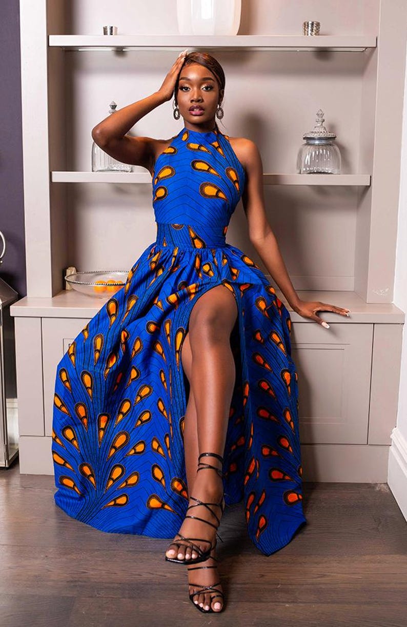 May include: A floor-length sleeveless dress in a vibrant blue fabric featuring a repeating orange and black teardrop pattern. The garment has a high halter neckline, a fitted waist, and a long skirt with a prominent front leg slit.