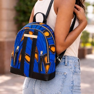 May include: A blue and orange patterned backpack with black leather detailing. The backpack features a gold zip and a gold bar on the front. The bag has black straps and a black base. The backpack is being worn.