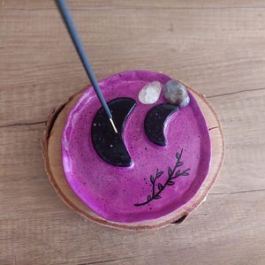 Peut inclure: Un porte-encens en céramique ronde, violette et mouchetée, orné de deux croissants de lune noirs et d'un motif de branche feuillue noire. Deux petites pierres lisses et translucides sont placées près des lunes et un bâton d'encens est inséré.