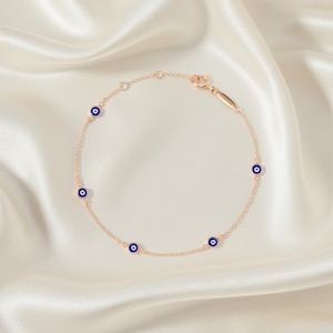 May include: A delicate rose gold chain bracelet featuring six small, round blue evil eye charms. The bracelet is displayed on a white satin surface, highlighting its fine details and elegant design. The bracelet's clasp is visible.