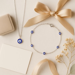 Puede incluir: Un conjunto de collar y pulsera de plata con abalorios de ojo turco azules y blancos. El collar tiene un solo colgante, mientras que la pulsera tiene varios abalorios. Una caja de regalo beige, una tarjeta en blanco y una cinta dorada.