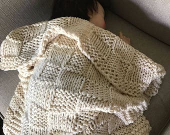 May include: A cream-colored, chunky knit blanket with a geometric pattern. The blanket is draped over a doll, creating a cozy and inviting scene. The blanket's texture and color suggest warmth and comfort.