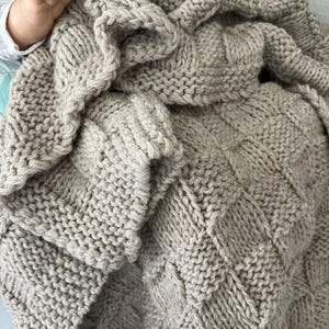 May include: A hand-knitted blanket in a light beige color featuring a textured basketweave pattern. The fabric is composed of thick yarn loops, creating a soft and chunky appearance with a repeating square grid design throughout the entire surface.