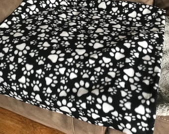Könnte beinhalten: Eine schwarze und weiße Fleece-Decke mit einem Pfotenabdruckmuster. Die Decke ist gefaltet und auf einem braunen Sofa platziert.