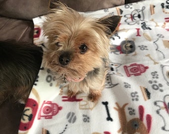 Puede incluir: Un pequeño Yorkshire Terrier marrón y blanco sentado en una manta blanca con un patrón de perro marrón, rojo y gris. La manta tiene las palabras "woof woof" impresas.