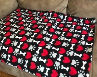 Könnte beinhalten: Eine schwarze Fleece-Hundebettdecke mit einem sich wiederholenden Muster aus roten Herzen, weißen Knochen und grauen Pfotenabdrücken.
