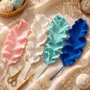 Puede incluir: Cuatro decoraciones de plumas de ganchillo en colores pastel: rosa, blanco, turquesa y azul. Las plumas tienen un diseño detallado y texturizado y están dispuestas sobre una superficie de tonos neutros. También se ven unas tijeras doradas y un ganchillo.