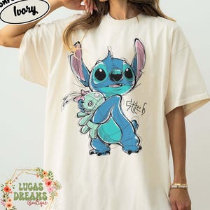 May include: An ivory-colored t-shirt featuring a graphic illustration of the blue character Stitch holding a small doll. The design includes the text Stitch in a stylized script. A Comfort Colors Ivory label is visible in the upper left corner.