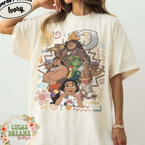 May include: An ivory short-sleeve t-shirt featuring a graphic print of characters from the movie Moana, including Moana, Maui, Pua, Heihei, and Gramma Tala. The design includes tribal-inspired patterns, a hook, and various colorful symbols.
