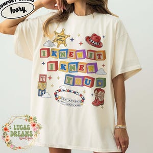 May include: An ivory short-sleeve t-shirt featuring a graphic print of colorful alphabet blocks spelling I KNEW IT I KNEW YOU. The design includes a sheriff badge with text INFINITY AND BEYOND, a cowboy hat, a beaded bracelet with text YOU'VE GOT A FRIEND IN ME, and cowboy boots.