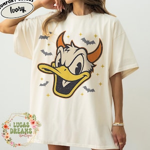 以下が含まれることがあります： アイボリー色のTシャツで、オレンジ色の悪魔の角が生えた漫画風のアヒルの顔が描かれています。デザインには小さな灰色のコウモリと黄色の四芒星が散りばめられています。左上にはCOMFORT COLORS IVORYというテキストが表示されています。