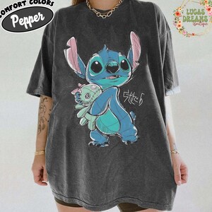 May include: A charcoal gray short-sleeve t-shirt featuring a graphic of the blue character Stitch holding a small doll. The design includes the text Stitch in a stylized script. The shirt has a vintage-washed appearance and a crew neckline.