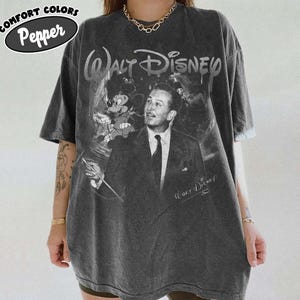 Peut inclure: Un t-shirt gris anthracite &agrave; manches courtes avec un graphisme de style vintage repr&eacute;sentant Walt Disney en costume-cravate accompagn&eacute; de Mickey Mouse. Le texte Walt Disney est imprim&eacute; en haut dans une police stylis&eacute;e, avec une signature sous le visuel. Le t-shirt est &eacute;tiquet&eacute; Comfort Colors Pepper.