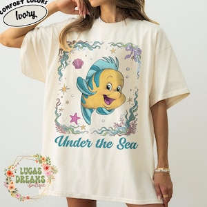May include: Ivory-colored t-shirt featuring a cartoon image of a yellow and blue fish with the text "Under the Sea." The design is framed with aquatic elements like seaweed, shells, and stars. The shirt is from Lucas Dreams Boutique.