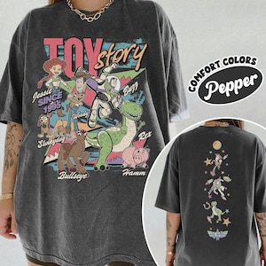 May include: Dark gray t-shirt featuring a graphic of Toy Story characters, including Woody, Buzz Lightyear, and Rex. The front of the shirt has the words "Toy Story" and "Jessie Since 1995". The back of the shirt has a vertical design of Toy Story characters.