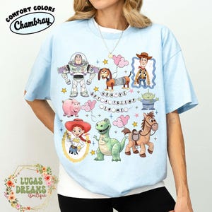 May include: Light blue t-shirt featuring cartoon characters including Buzz Lightyear, Woody, and Rex. The shirt has the phrase "YOU'VE GOT A FRIEND IN ME" and the Comfort Colors and Chambray logos. The shirt is from Lucas Dreams Boutique.