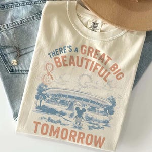 May include: A cream-colored short-sleeve t-shirt featuring a graphic print with the text "THERE'S A GREAT BIG BEAUTIFUL TOMORROW." The design includes a blue illustration of a circular building, a sun, clouds, and a small mouse character.
