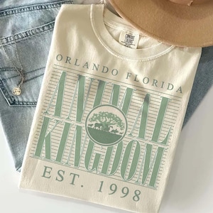 May include: Cream-colored t-shirt with "Orlando Florida Animal Kingdom" printed in green. The design includes a tree and animal silhouettes, with "EST. 1998" below. The shirt is folded on top of blue jeans.