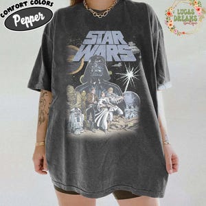 Pode incluir: T-shirt cinza escuro com estampa gráfica de Star Wars. A camisa apresenta o logotipo de Star Wars e um gráfico de Darth Vader e outros personagens. A camisa tem as palavras "Comfort Colors Pepper" em um oval preto no canto superior esquerdo.