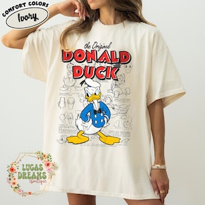 May include: Ivory-colored t-shirt featuring a graphic of Donald Duck in a blue jacket and bow tie. The text "The Original DONALD DUCK" is in red and black. The shirt has a relaxed fit and short sleeves.
