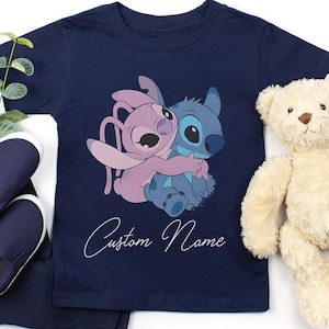 May include: Navy blue t-shirt featuring a graphic of two cartoon characters hugging, one pink and one blue. The text "Custom Name" is written in cursive below the graphic. A teddy bear and a pair of blue shoes are also in the image.