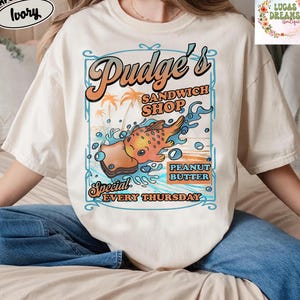 Pu&ograve; includere: T-shirt Comfort Colors color avorio con grafica per "Pudge's Sandwich Shop". Il design include un sandwich di pesce dei cartoni animati, palme e la scritta "Peanut Butter Special Every Thursday."