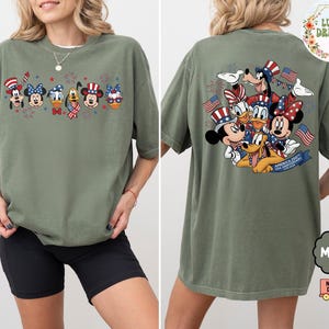 以下が含まれることがあります： モスグリーンのTシャツ。愛国的な漫画のデザインが特徴です。前面には、お祝いの帽子と花火をかぶった6人のキャラクターが描かれています。背面には、アメリカの国旗を持ったキャラクターのグループと「America 250th USA Anniversary」の文字が描かれています。
