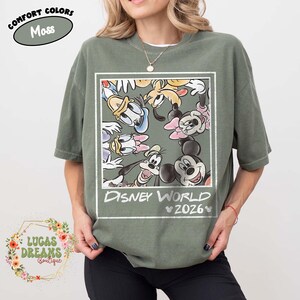 Op de afbeelding: Mosgroen T-shirt met een Disney World-afbeelding met Mickey Mouse, Minnie Mouse, Donald Duck en Goofy. De tekst "Disney World 2026" staat onder de karakters. Het shirt heeft korte mouwen en een ronde hals.