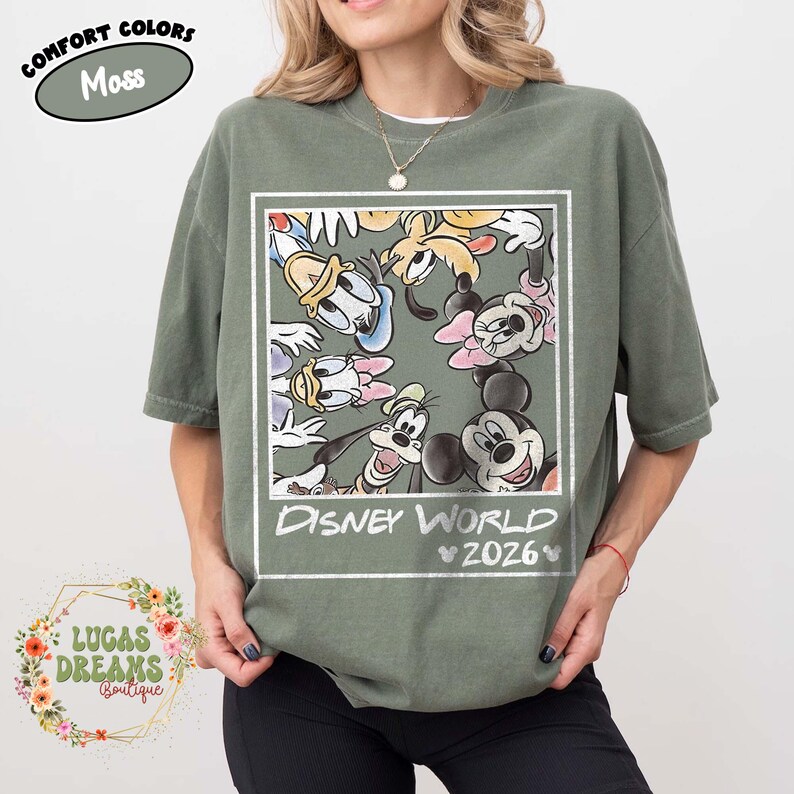 Op de afbeelding: Mosgroen T-shirt met een Disney World-afbeelding met Mickey Mouse, Minnie Mouse, Donald Duck en Goofy. De tekst "Disney World 2026" staat onder de karakters. Het shirt heeft korte mouwen en een ronde hals.