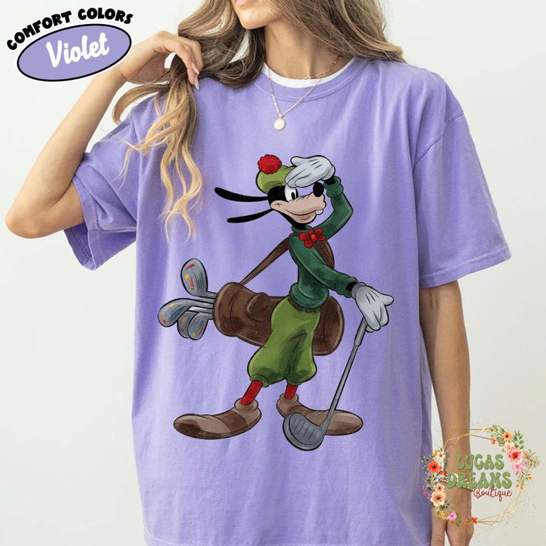 May include: Violet Comfort Colors t-shirt featuring a cartoon Goofy character dressed in golf attire. Goofy is holding a golf club and a golf bag. The shirt has the words "Comfort Colors" and "Violet" in a circle.