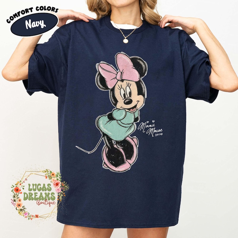 May include: A navy blue short-sleeve t-shirt featuring a graphic of Minnie Mouse wearing a light pink bow and a teal dress. The text "Minnie Mouse xoxo" is printed in white script next to the character. The shirt has a crew neckline.