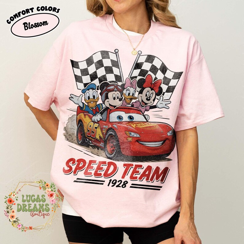 Op de afbeelding: Zachtroze T-shirt met een afbeelding van Disney-personages Mickey Mouse, Donald Duck, Daisy Duck en Minnie Mouse in een rode raceauto met de tekst "SPEED TEAM 1928". Twee geblokte vlaggen op de achtergrond. Het shirt heeft de tekst "COMFORT COLORS Blossom".