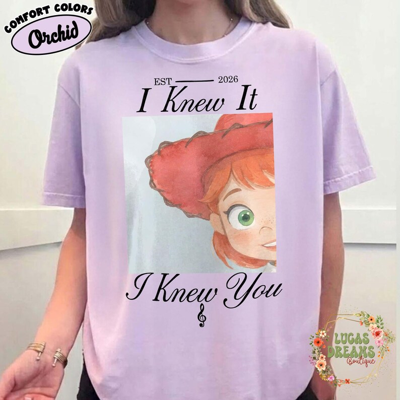 May include: A light purple short-sleeve t-shirt featuring a graphic of a character with red hair and a red hat. The text reads EST 2026, I Knew It, and I Knew You with a musical note symbol. The shirt has a crew neckline and a relaxed fit.