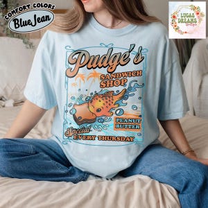 Pu&ograve; includere: T-shirt azzurra con grafica retr&ograve; per "Pudge's Sandwich Shop". Il design presenta un pesce dei cartoni animati, un panino con burro di arachidi e la scritta "Special Every Thursday". La maglietta ha anche la scritta "Comfort Colors Blue Jean."