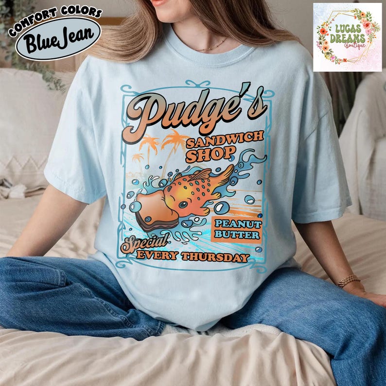 Pu&ograve; includere: T-shirt azzurra con grafica retr&ograve; per "Pudge's Sandwich Shop". Il design presenta un pesce dei cartoni animati, un panino con burro di arachidi e la scritta "Special Every Thursday". La maglietta ha anche la scritta "Comfort Colors Blue Jean."
