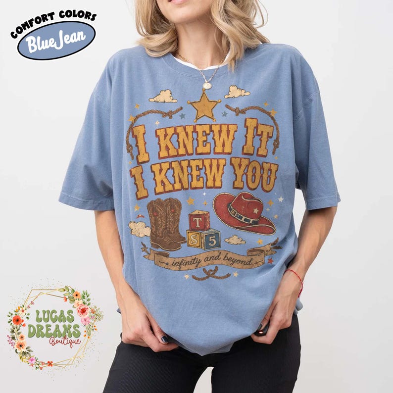 May include: A blue short-sleeve t-shirt featuring a graphic print with the text I KNEW IT I KNEW YOU and infinity and beyond. The design includes a yellow star, brown cowboy boots, a red cowboy hat, and two alphabet blocks labelled T and 5.