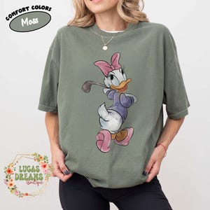 May include: Moss green t-shirt featuring a cartoon Daisy Duck playing golf. Daisy Duck is wearing a purple shirt, pink bow, and pink shoes. The shirt has the text "COMFORT COLORS" and "Moss" in a green oval. The shop name "LUCAS DREAMS BOUTIQUE" is also visible.