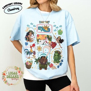 May include: Pale blue t-shirt with a vibrant Disney-themed graphic. The design showcases characters like Moana, Maui, and Hei Hei. The word "Disney" is at the top. The shirt also features the Comfort Colors and Lucas Dreams Boutique logos.