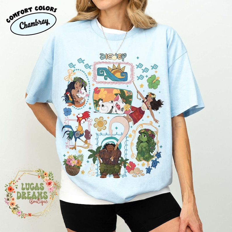 May include: Pale blue t-shirt with a vibrant Disney-themed graphic. The design showcases characters like Moana, Maui, and Hei Hei. The word "Disney" is at the top. The shirt also features the Comfort Colors and Lucas Dreams Boutique logos.